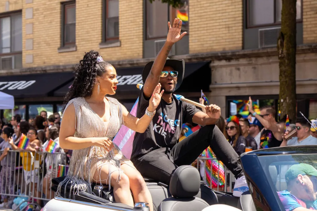 Precious Brady-Davis et Myles Brady-Davis sont assis à l'arrière d'une décapotable et saluent la foule lors de la Chicago Pride Parade. Les drapeaux de la fierté sont visibles dans la foule derrière eux.
